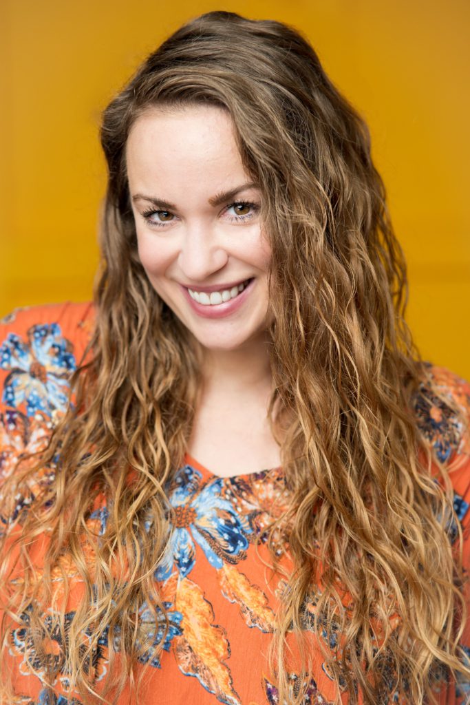 Jillian has light skin, warm brown eyes, and long wavy brown hair. She smiles and wears an orange and blue floral top.